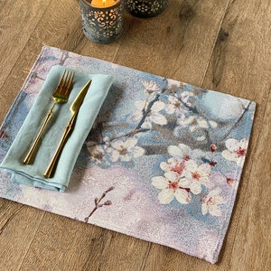 May include: A blue and white floral placemat with a light blue napkin and gold silverware on top. The placemat is on a wooden table with two candle holders.