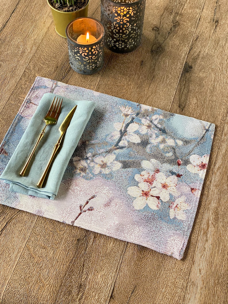 May include: A blue and white floral placemat with a light blue napkin and gold silverware on top. The placemat is on a wooden table with two candle holders.