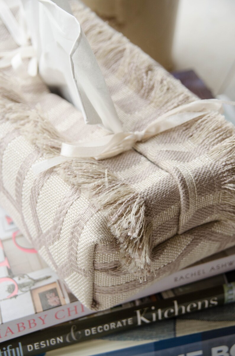 Peut inclure: Un couvre-bo&icirc;te &agrave; mouchoirs beige avec un bo&icirc;te &agrave; mouchoirs blanche &agrave; l'int&eacute;rieur. Le couvre-bo&icirc;te est orn&eacute; d'une frange et d'un ruban blanc nou&eacute; autour. Le bo&icirc;te &agrave; mouchoirs est pos&eacute; sur une pile de livres.