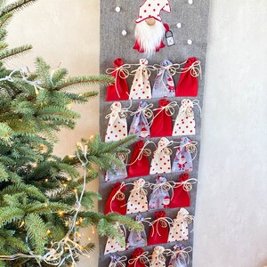 Puede incluir: Un calendario de adviento de fieltro gris con 24 bolsitas de tela roja y blanca. Cada bolsita tiene una cuerda blanca y una campana dorada. Un gnomo rojo y blanco con barba blanca est&aacute; en la parte superior del calendario.