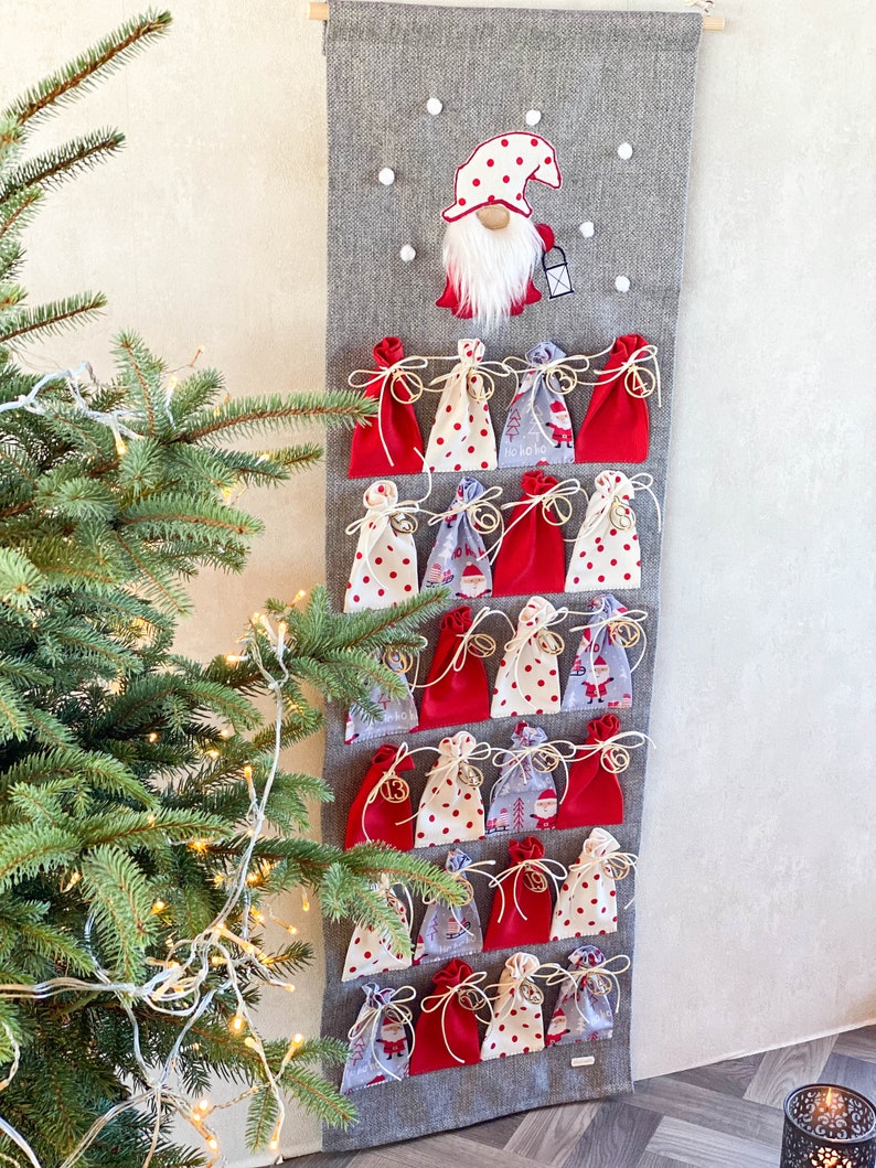 Puede incluir: Un calendario de adviento de fieltro gris con 24 bolsitas de tela roja y blanca. Cada bolsita tiene una cuerda blanca y una campana dorada. Un gnomo rojo y blanco con barba blanca est&aacute; en la parte superior del calendario.
