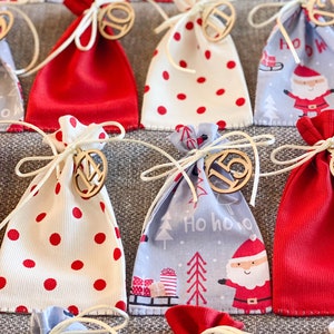 Puede incluir: Un calendario de adviento navide&ntilde;o con 24 peque&ntilde;as bolsas de tela. Las bolsas est&aacute;n decoradas con lunares rojos y blancos, un patr&oacute;n de Pap&aacute; Noel azul y blanco y una tela roja lisa. Cada bolsa tiene una etiqueta de madera numerada.