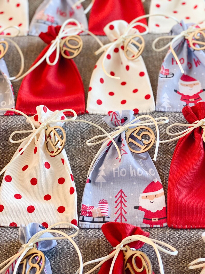 Puede incluir: Un calendario de adviento navide&ntilde;o con 24 peque&ntilde;as bolsas de tela. Las bolsas est&aacute;n decoradas con lunares rojos y blancos, un patr&oacute;n de Pap&aacute; Noel azul y blanco y una tela roja lisa. Cada bolsa tiene una etiqueta de madera numerada.