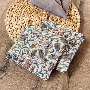 Könnte beinhalten: Zwei Topflappen mit floralem Muster mit Vögeln und Blumen in den Farben Blau, Grün, Braun und Weiß. Die Topflappen liegen auf einer geflochtenen Stroh-Unterlage.