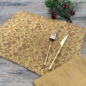 Puede incluir: Un mantel individual dorado con un estampado floral, un tenedor y un cuchillo dorados y una servilleta a juego. El mantel individual es rectangular y tiene un diseño detallado y repetitivo. La cubertería tiene un diseño sencillo y elegante.