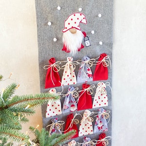 Puede incluir: Un calendario de adviento de fieltro gris con un gnomo rojo y blanco en la parte superior. El calendario tiene 24 bolsillos de tela roja, blanca y azul. Cada bolsillo tiene una cuerda dorada y una peque&ntilde;a campana dorada.