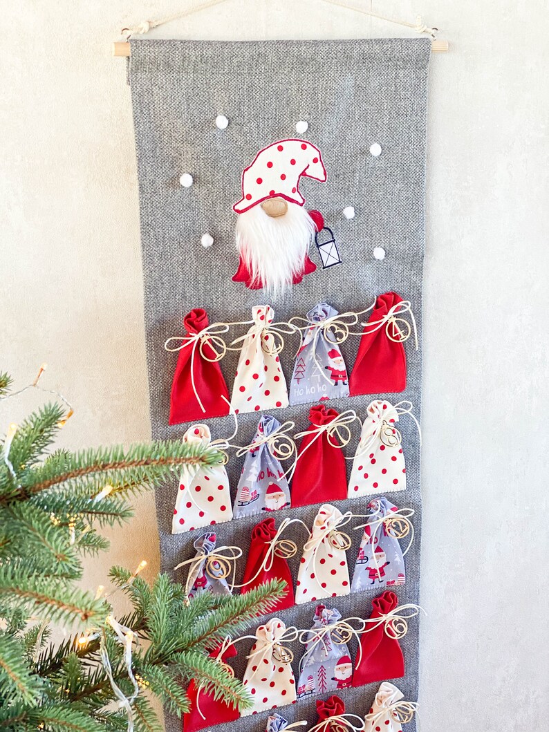 Puede incluir: Un calendario de adviento de fieltro gris con un gnomo rojo y blanco en la parte superior. El calendario tiene 24 bolsillos de tela roja, blanca y azul. Cada bolsillo tiene una cuerda dorada y una peque&ntilde;a campana dorada.