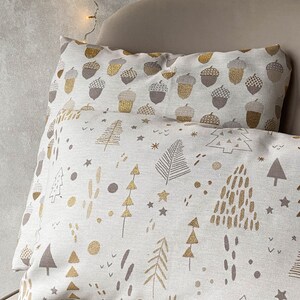 May include: Two decorative throw pillows with a white background and gold and brown patterns. The top pillow has a pattern of acorns, while the bottom pillow has a pattern of trees and stars.