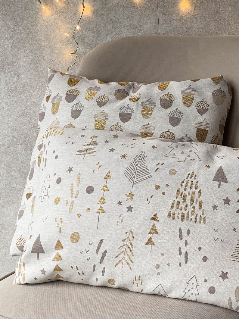 May include: Two decorative throw pillows with a white background and gold and brown patterns. The top pillow has a pattern of acorns, while the bottom pillow has a pattern of trees and stars.