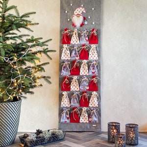 Puede incluir: Un calendario de adviento de tela gris con 24 bolsas de tela roja y blanca. Un gnomo con un gorro rojo y barba blanca est&aacute; en la parte superior del calendario.