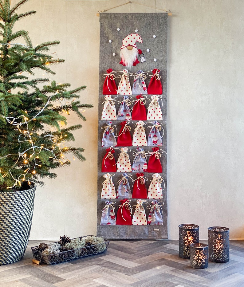 Puede incluir: Un calendario de adviento de tela gris con 24 bolsas de tela roja y blanca. Un gnomo con un gorro rojo y barba blanca est&aacute; en la parte superior del calendario.