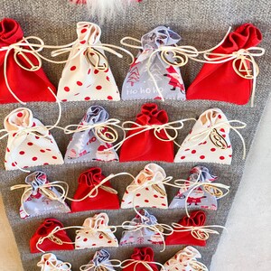 Puede incluir: Un calendario de adviento de fieltro gris con 24 bolsas de tela roja, blanca y gris numeradas. Las bolsas est&aacute;n atadas con cuerda y tienen n&uacute;meros de madera.