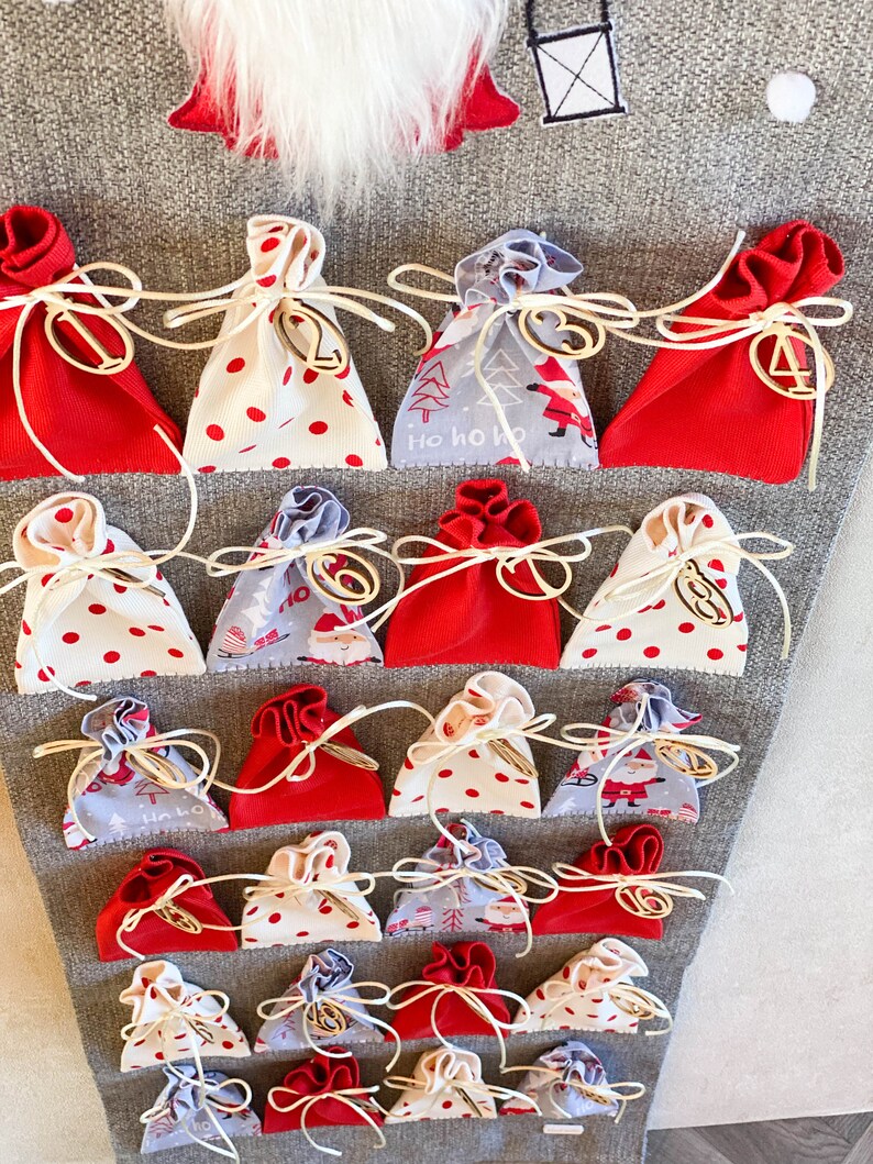 Puede incluir: Un calendario de adviento de fieltro gris con 24 bolsas de tela roja, blanca y gris numeradas. Las bolsas est&aacute;n atadas con cuerda y tienen n&uacute;meros de madera.