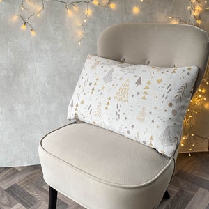 May include: A beige armchair with black legs, featuring a white throw pillow with a gold and brown geometric pattern of trees and stars. The chair is set against a gray wall with string lights.