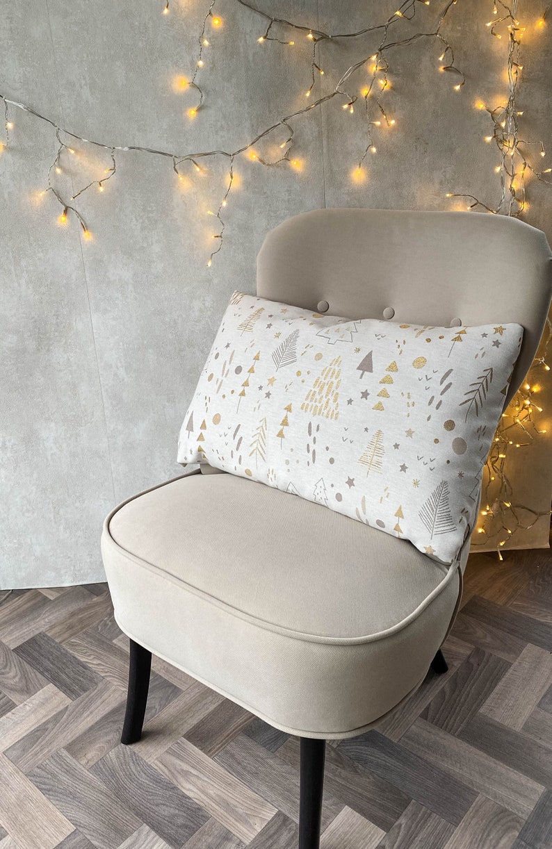 May include: A beige armchair with black legs, featuring a white throw pillow with a gold and brown geometric pattern of trees and stars. The chair is set against a gray wall with string lights.