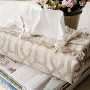 Peut inclure: Un couvre-bo&icirc;te &agrave; mouchoirs beige avec un motif g&eacute;om&eacute;trique et une bordure en franges. Le couvre-bo&icirc;te est nou&eacute; avec un ruban blanc et repose sur une pile de livres.