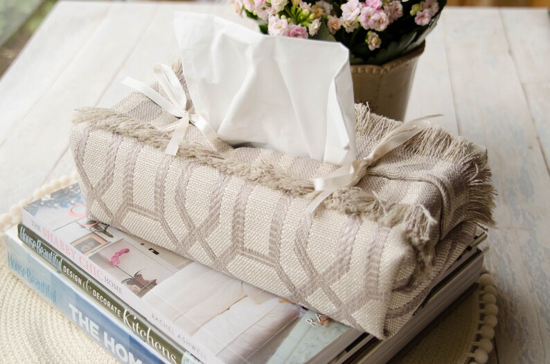 Peut inclure: Un couvre-bo&icirc;te &agrave; mouchoirs beige avec un motif g&eacute;om&eacute;trique et une bordure en franges. Le couvre-bo&icirc;te est nou&eacute; avec un ruban blanc et repose sur une pile de livres.