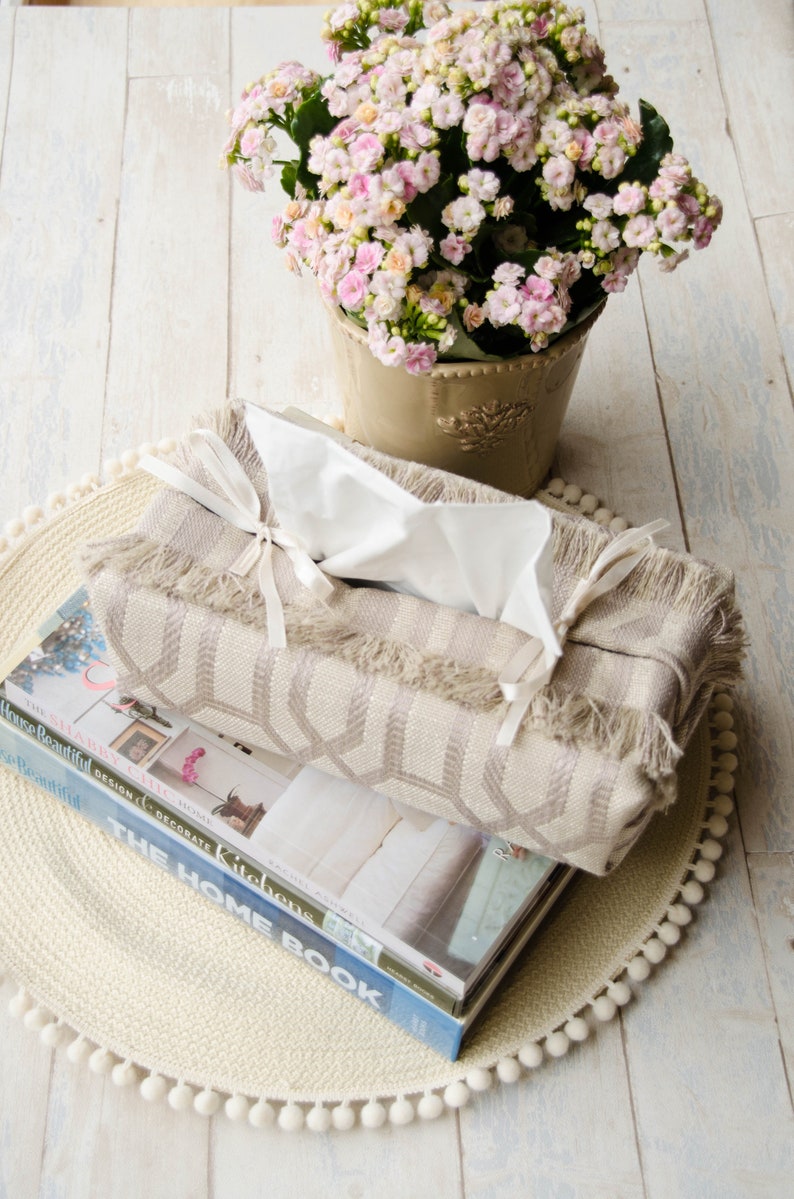Peut inclure: Un couvre-bo&icirc;te &agrave; mouchoirs beige avec une bordure en franges et des rubans blancs est pos&eacute; sur une pile de livres avec un tapis &agrave; pompons blancs. Une plante en pot avec des fleurs roses est en arri&egrave;re-plan.