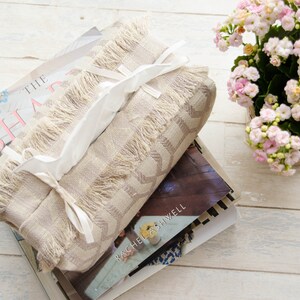 Peut inclure: Un couvre-bo&icirc;te &agrave; mouchoirs beige avec une bordure en franges et des liens en ruban blanc. Le couvre-bo&icirc;te est pos&eacute; sur une pile de livres. Le livre du dessus porte le titre "The Shape" et le livre du dessous porte le titre "Rachel Ashwell".