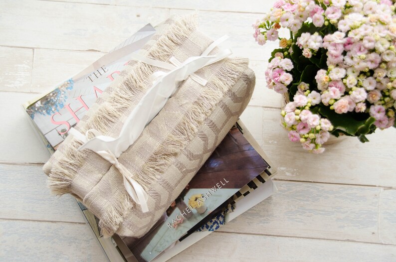 Peut inclure: Un couvre-bo&icirc;te &agrave; mouchoirs beige avec une bordure en franges et des liens en ruban blanc. Le couvre-bo&icirc;te est pos&eacute; sur une pile de livres. Le livre du dessus porte le titre "The Shape" et le livre du dessous porte le titre "Rachel Ashwell".