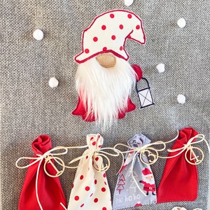 Puede incluir: Un calendario de adviento de fieltro gris con bolsas de tela roja y blanca. Cada bolsa tiene una etiqueta de madera con un n&uacute;mero. Un gnomo con un gorro rojo y barba blanca est&aacute; en la parte superior del calendario.