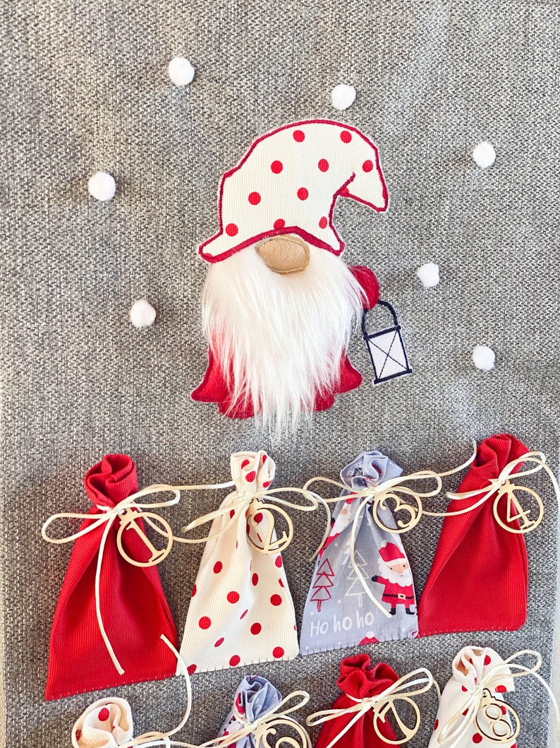 Puede incluir: Un calendario de adviento de fieltro gris con bolsas de tela roja y blanca. Cada bolsa tiene una etiqueta de madera con un n&uacute;mero. Un gnomo con un gorro rojo y barba blanca est&aacute; en la parte superior del calendario.