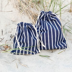 May include: Two drawstring pouches made of navy blue fabric with vertical white stripes. Each bag features a gathered top closure secured by thick, off-white cotton cords. The pouches are of different sizes, with one being noticeably larger.
