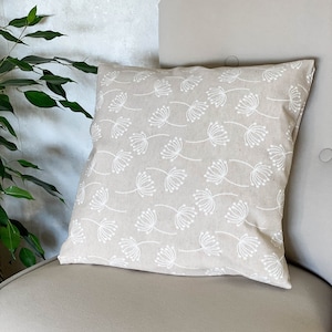 May include: A beige throw pillow with a white floral pattern. The pillow is sitting on a beige armchair.