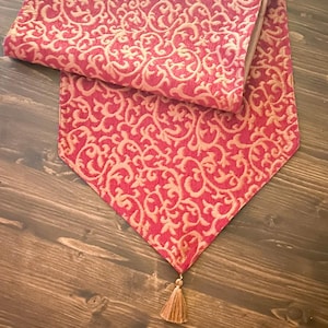 Op de afbeelding: Een rode tafelloper met een crèmekleurig, sierlijk, wervelend patroon. De loper is aan de bovenkant opgevouwen en heeft aan de onderkant een kwast. De loper is gemaakt van een textuurstof.