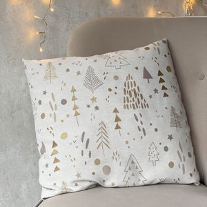 May include: A white throw pillow with a gold and brown Christmas tree pattern. The pillow is sitting on a light brown couch.