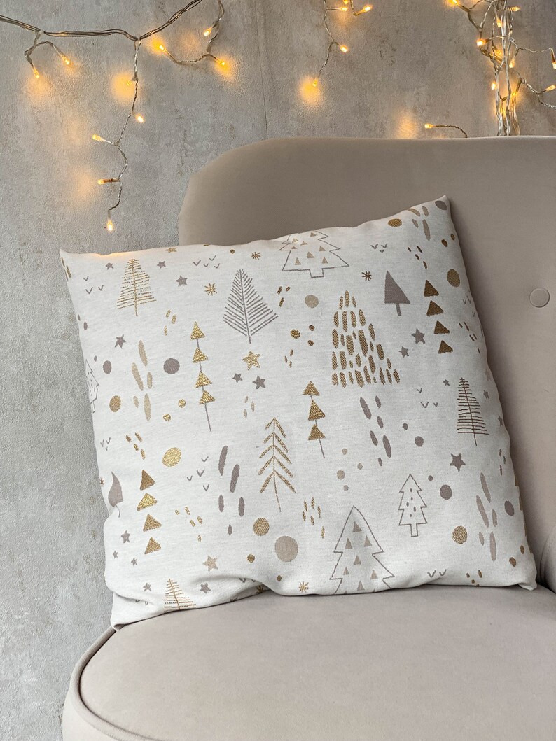 May include: A white throw pillow with a gold and brown Christmas tree pattern. The pillow is sitting on a light brown couch.