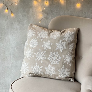 May include: A beige square pillow with a white snowflake pattern rests on a beige armchair. The pillow's design features various sizes of detailed snowflakes. The background includes a gray wall and a string of warm white fairy lights.
