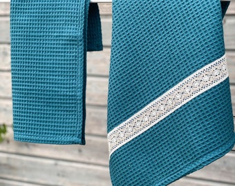 May include: Two teal waffle weave kitchen towels hanging on a clothesline. One towel has a white lace trim.