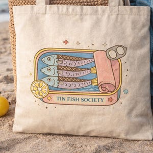 Sardine tote bag with Tin Fish Society design on beach sand with lemons and coastal aesthetic styling