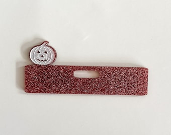 Puede incluir: Una etiqueta de nombre de Halloween de purpurina roja con un recorte de calabaza blanca. La calabaza tiene una cara tallada.