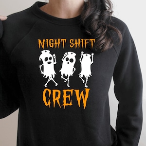 Op de afbeelding: Zwart sweatshirt met een witte afbeelding van drie geesten die verpleegstersmutsen dragen en de tekst "NIGHT SHIFT CREW" in oranje.