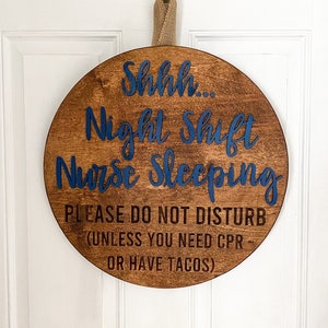 May include: A round wooden sign with the text "Shhh... Night Shift Nurse Sleeping. Please do not disturb (unless you need CPR or have tacos)" in blue lettering.