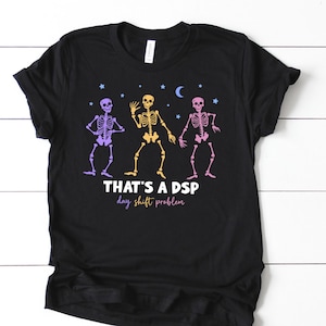 May include: Black t-shirt featuring a graphic of three dancing skeletons in purple, yellow, and pink. The text "THAT'S A DSP day shift problem" is printed below the skeletons. The t-shirt is a crew neck.