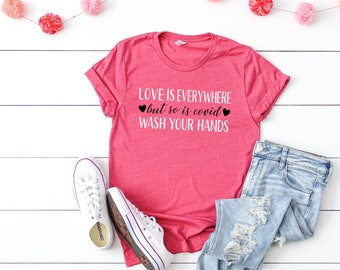 May include: A pink t-shirt with the text "LOVE IS EVERYWHERE but so is covid WASH YOUR HANDS" in white with black hearts on a white background. The shirt is laid out on a white wooden surface with a string of pink pom-poms.