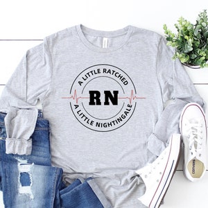 Op de afbeelding: Een grijs longsleeve shirt met een zwarte cirkelvormige afbeelding met de tekst "A Little Ratched, A Little Nightingale" met een rode hartslaglijn en de letters "RN" in het midden.