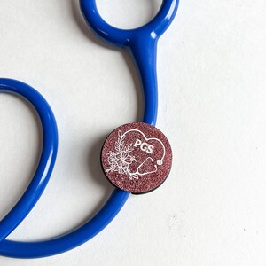 May include: A stethoscope with a pink glitter charm attached. The charm has a white heart with the text "PGS" inside and a floral design.