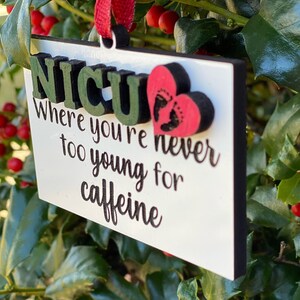 K&ouml;nnte beinhalten: Wei&szlig;er Holzanh&auml;nger mit dem Text "NICU Where you're never too young for caffeine" und einem roten Herz mit einem Fu&szlig;abdruck-Design.