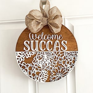 Peut inclure: Un porte-manteau rond en bois avec un n&oelig;ud en toile de jute. Le porte-manteau a un motif d&eacute;coup&eacute; blanc de succulentes et les mots "Welcome Succas" en lettres blanches.
