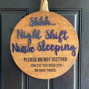 May include: A round wooden sign with the text "Shhh... Night Shift Nurse Sleeping" in blue lettering. The sign also includes the text "PLEASE DO NOT DISTURB (UNLESS YOU NEED CPR - OR HAVE TACOS)" in black lettering.