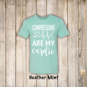 May include: A light blue t-shirt with the text "COMPRESSIONS ARE MY cardio" in white. A stethoscope and a heart beat graphic are above the text. The shirt is on a wood background.