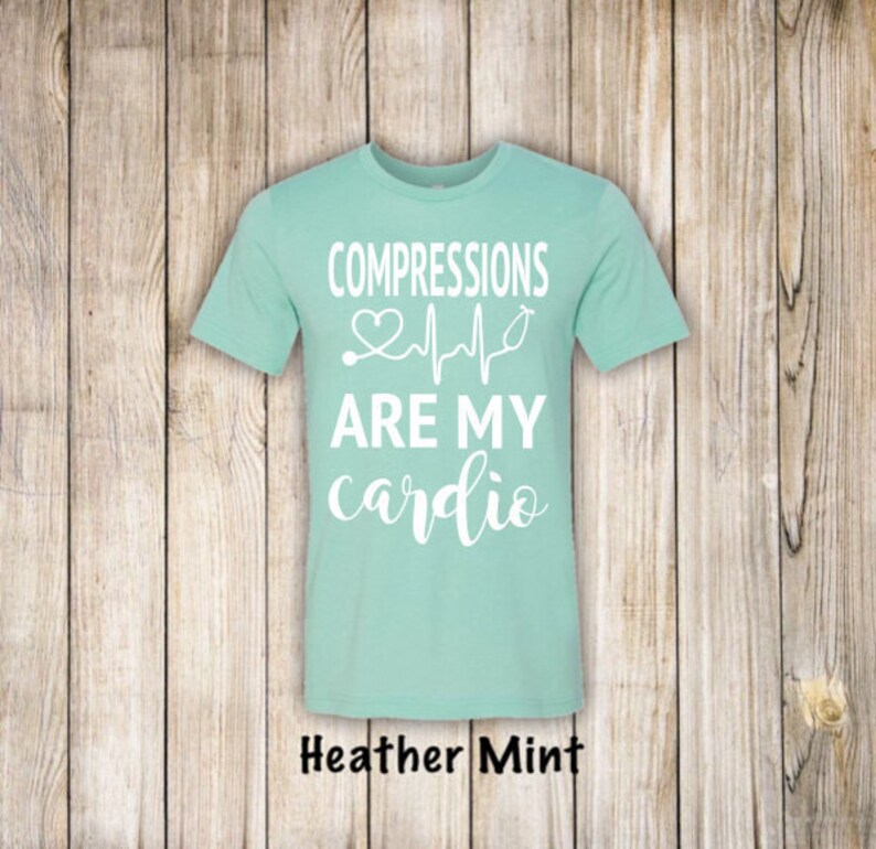 May include: A light blue t-shirt with the text "COMPRESSIONS ARE MY cardio" in white. A stethoscope and a heart beat graphic are above the text. The shirt is on a wood background.