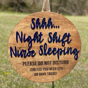 May include: A round wooden sign with the text "Shhh... Night Shift Nurse Sleeping PLEASE DO NOT DISTURB (UNLESS YOU NEED CPR - OR HAVE TACOS)" in blue and black lettering.