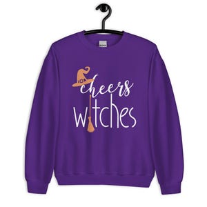 Op de afbeelding: Een paars sweatshirt met lange mouwen en ronde hals met de witte tekst "cheers witches" op de voorzijde. Het ontwerp bevat een lichtoranje heksenhoed boven het woord "cheers" en een lichtoranje bezemsteel die de letter "i" vervangt.