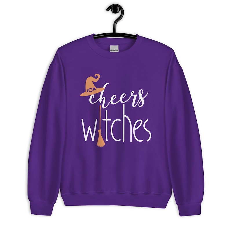 Op de afbeelding: Een paars sweatshirt met lange mouwen en ronde hals met de witte tekst "cheers witches" op de voorzijde. Het ontwerp bevat een lichtoranje heksenhoed boven het woord "cheers" en een lichtoranje bezemsteel die de letter "i" vervangt.
