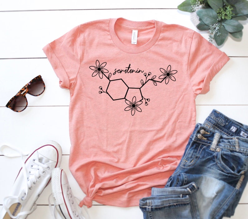 Puede incluir: Una camiseta rosa con un gr&aacute;fico negro de una mol&eacute;cula de serotonina con flores. El texto "serotonin" est&aacute; escrito en el gr&aacute;fico.